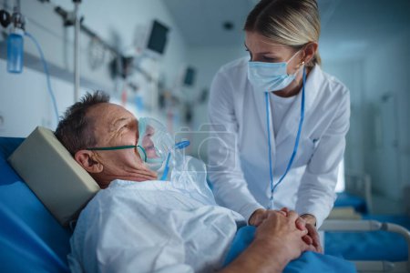 Junge Ärztin spricht ältere Patientin mit Sauerstoffmaske im Krankenhauszimmer an.