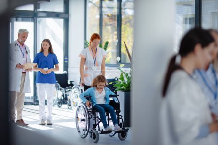 Téléchargez les photos : Jeune infirmière poussant une petite fille en fauteuil roulant dans un couloir d'hôpital. - en image libre de droit