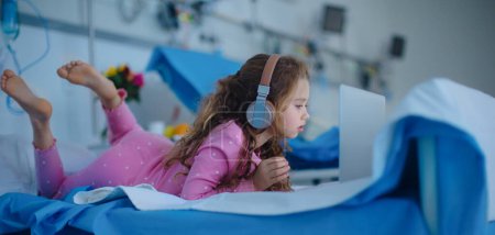 Photo for Little sick girl lying alone at hospital bed and using a laptop. - Royalty Free Image
