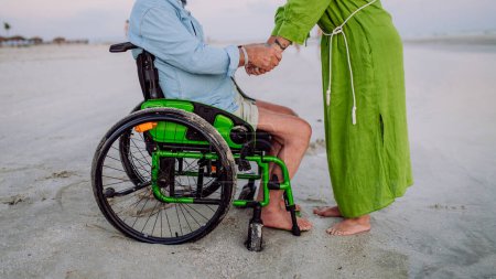 Téléchargez les photos : Senior man on wheelchair enjoying together time with his wife at the sea, low section. - en image libre de droit
