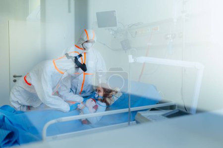 Foto de Niña con una enfermedad infecciosa acostada en la habitación del hospital, médicos con coberturas protectoras examinándola. - Imagen libre de derechos