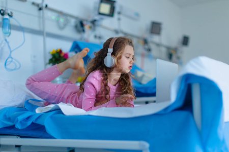 Photo for Little sick girl lying alone at hospital bed and using a laptop. - Royalty Free Image