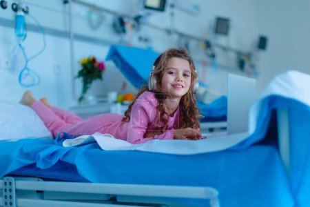 Téléchargez les photos : Petite fille malade couchée seule au lit d'hôpital et utilisant un ordinateur portable. - en image libre de droit