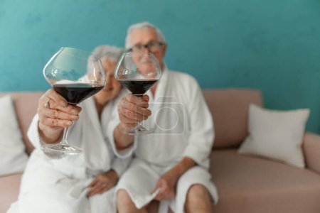 Photo for Happy senior couple sitting together in the bathrobe on sofa with glass of wine, having nice time at home. - Royalty Free Image