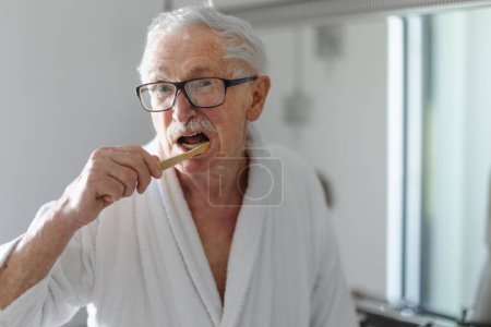 Téléchargez les photos : Senior homme nettoyer ses dents avec une brosse en bois dans la salle de bain, mode de vie durable. - en image libre de droit