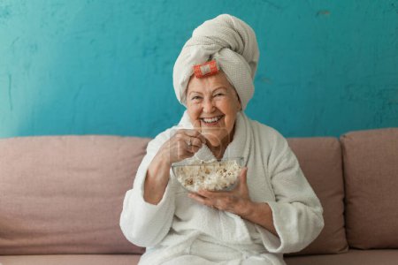 Foto de Happy senior woman sitting at a sofa in bathrobes and watching TV with popcorn. - Imagen libre de derechos