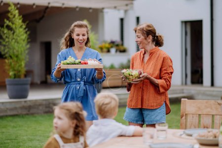 Téléchargez les photos : Femmes heureuses apportant une salade et des hamburgers à la fête de jardin multi-génération en été. - en image libre de droit