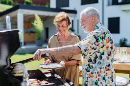 Photo for Senior man grilled outdoor at garden, giving his wife fresh hamburger. - Royalty Free Image