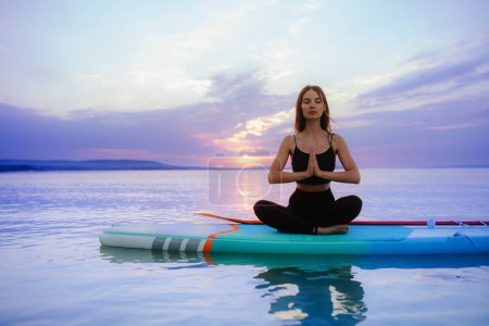 Téléchargez les photos : A young surfer girl meditating on surfboard on the lake at sunrise. - en image libre de droit