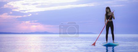 Photo for A young beautiful girl surfer paddling on surfboard on the lake at sunrise - Royalty Free Image