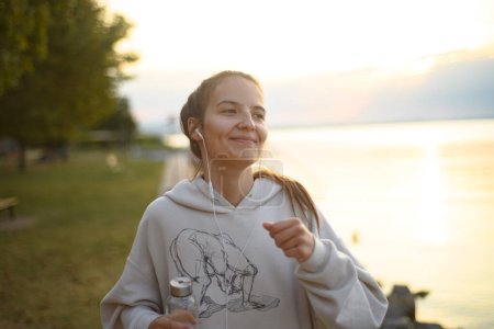 Photo for A young beautiful sportive girl listening to music and jogging at sunrise by the lake. - Royalty Free Image