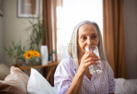 Photo for Portrait of senior woman sitting on the bed in a pajamas and drinking water. - Royalty Free Image