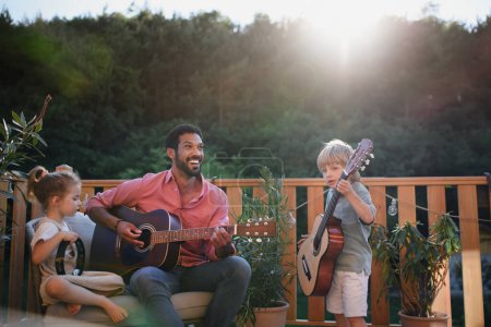 Foto de Un joven afroamericano disfruta tocando la guitarra con niños pequeños acompañándolo con instrumentos en el patio en el jardín en verano. - Imagen libre de derechos
