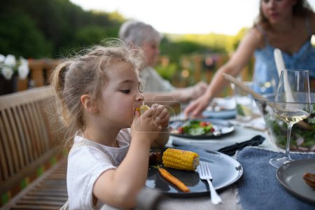 Téléchargez les photos : Une famille qui mange au barbecue dîner sur le patio, une petite fille qui mange du maïs rôti et en profite. - en image libre de droit