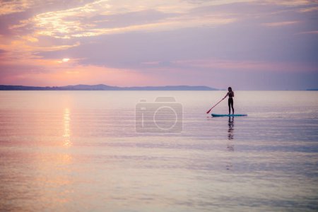 Foto de A young beautiful girl surfer paddling on surfboard on the lake at sunrise - Imagen libre de derechos
