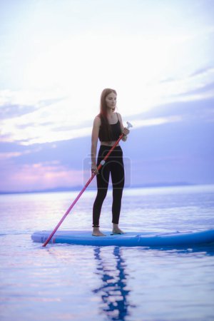 Téléchargez les photos : A young beautiful girl surfer paddling on surfboard on the lake at sunrise - en image libre de droit