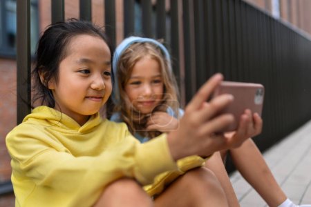 Téléchargez les photos : Petites filles avec capuche assise sur le trottoir et faisant défiler un smartphone. - en image libre de droit