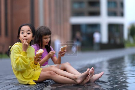 Téléchargez les photos : Two girls sitting, having legs in a fountain, enjoying ice cream and summer day. - en image libre de droit