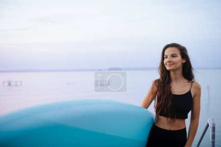 Téléchargez les photos : A young beautiful sportive girl with paddleboard on the lake at sunrise - en image libre de droit