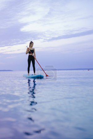 Téléchargez les photos : A young beautiful girl surfer paddling on surfboard on the lake at sunrise - en image libre de droit