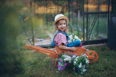 Photo for Little girl playing with wooden wheelbarrow full of flowers in the garden. - Royalty Free Image