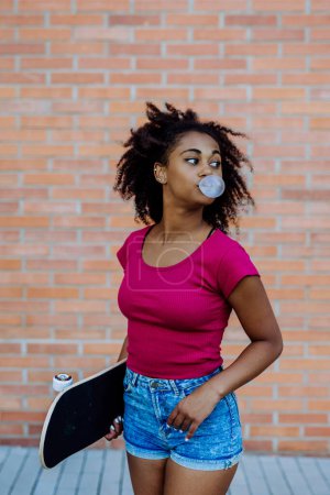 Téléchargez les photos : Adolescente multiraciale avecchewing-gum et planche à roulettes, marchant en ville pendant une journée d'été. - en image libre de droit