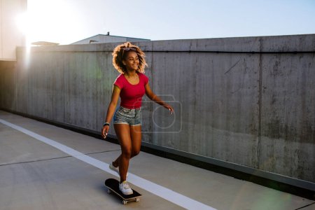 Téléchargez les photos : Multiracial teenage girl riding skateboard in front of concrete wall, balancing. Side view. - en image libre de droit