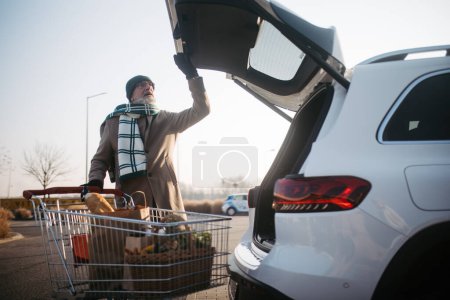 Téléchargez les photos : Senior homme donnant son achat dans le coffre de la voiture électrique. - en image libre de droit