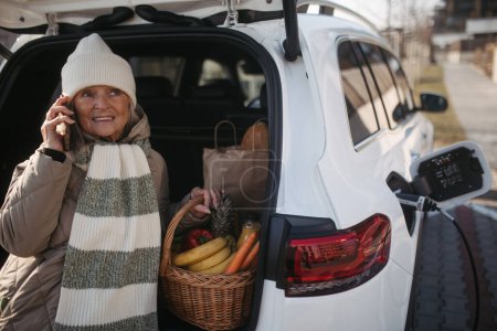 Foto de Mujer mayor sentada en el maletero del coche con compra de comestibles y esperando a cargar su coche eléctrico. - Imagen libre de derechos