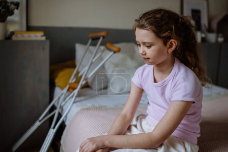 Photo for Little unhappy girl with broken leg sitting on a bed in her room. - Royalty Free Image