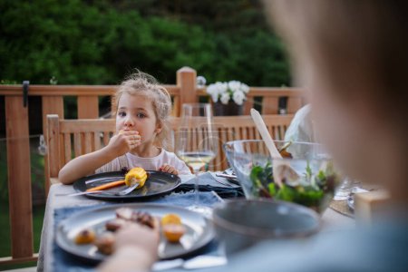 Photo for A family eating at barbecue party dinner on patio, little girl eating roasted corn and enjoying it. - Royalty Free Image