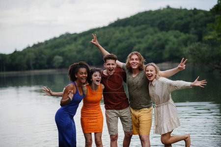 Foto de Un grupo multirracial de jóvenes amigos tomados de la mano y de pie en el lago en verano. - Imagen libre de derechos