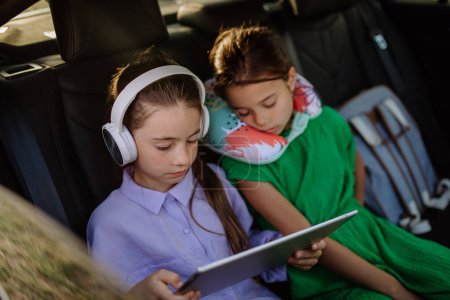 Foto de Chica little mirando en la tableta digital y escuchando música, su hermana se duerme durante el viaje familiar en coche. - Imagen libre de derechos