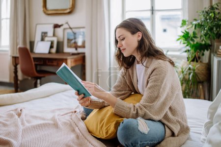 Téléchargez les photos : Jeune femme jouissant de temps libre dans son appartement, la lecture d'un livre. - en image libre de droit