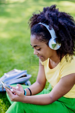 Téléchargez les photos : Multiracial girl sitting in a grass and enjoying music in headphones, side view. - en image libre de droit