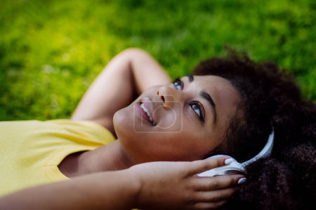 Photo for Multiracial girl lying down in a grass and enjoying music in headphones, side view. - Royalty Free Image