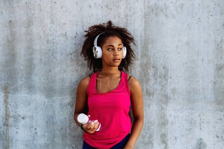 Photo for Young multiracial girl in sportswear resting after jogging in city, drinking water from a sustainable bottle and listening music in headphones. - Royalty Free Image