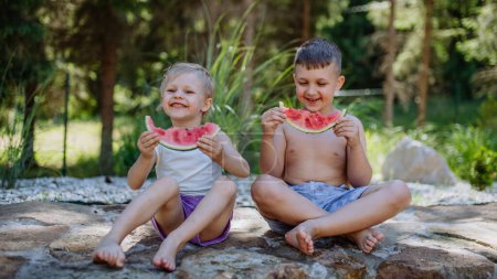 Photo for Little chidren sitting near a lake and eating watermelon on hot sunny day during summer vacation. - Royalty Free Image