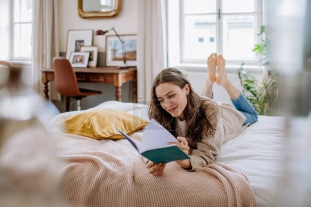 Photo for Young woman enjoying leisure time in her apartment, reading a book. - Royalty Free Image