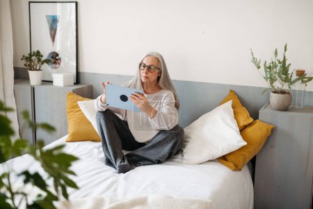 Photo for Senior woman sitting on a bed with a digital tablet. - Royalty Free Image