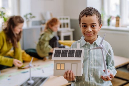 Téléchargez les photos : Petit garçon posant avec un modèle d'éolienne et modèle de maison avec système solaire pendant la leçon d'école. - en image libre de droit