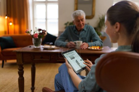 Téléchargez les photos : Aide-soignant lisant un journal en ligne dans une tablette numérique pendant qu'il prend soin d'un aîné chez lui. - en image libre de droit