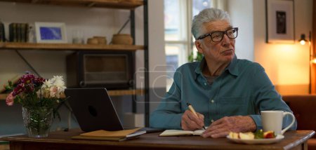Foto de Hombre mayor escribiendo notas en su diario, con computadora abierta. - Imagen libre de derechos