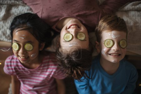 Foto de Vista superior de tres amigos felices acostados en la cama con máscara de belleza en la cara. - Imagen libre de derechos