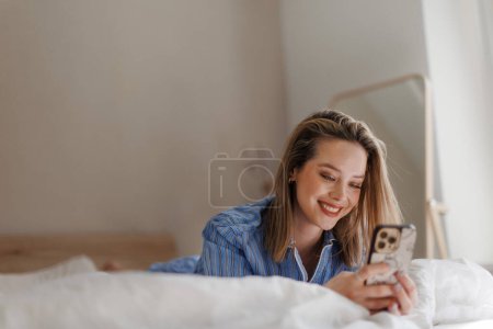 Téléchargez les photos : Jeune femme allongée sur son lit et faisant défiler un smartphone. - en image libre de droit
