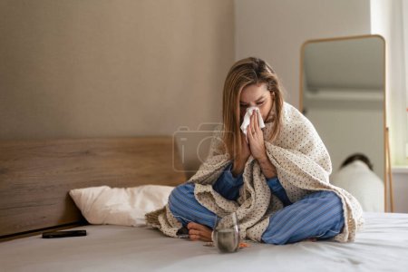 Kranke Frau sitzt mit Decke auf einem Bett.