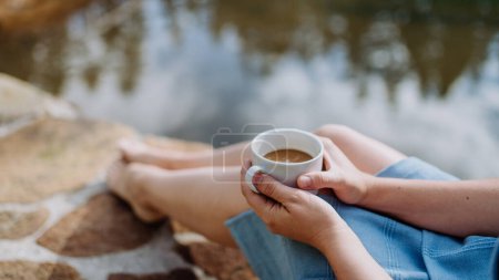 Foto de Primer plano de la mujer sentada cerca del lago y disfrutando de una taza de café. Vista de ángulo alto. - Imagen libre de derechos