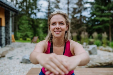 Téléchargez les photos : Jeune femme heureuse faisant de l'exercice, s'étendant sur un patio de chalet de montagne entouré par la nature. - en image libre de droit
