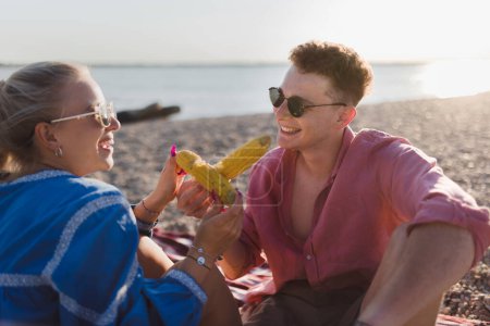 Foto de A happy young couple dating together in beach, sitting on a blanket and eating corn. Enjoying holiday time together. - Imagen libre de derechos
