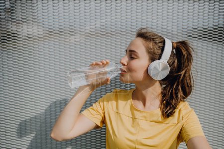 Photo for Young woman drinking water and listening music during jogging in a city, healthy lifestyle and sport concept. - Royalty Free Image
